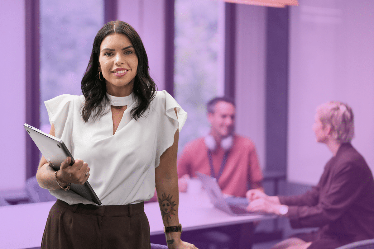 A woman standing in a modern office holding a laptop. In the background, two colleagues are sitting at a table having a discussion.